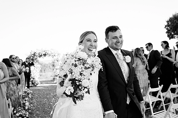 Recessional moment as bride and groom walk up the aisle, bride laughing with bouquet, guests beside white chairs on a bright lawn