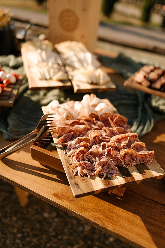 Wedding charcuterie board with cocktail hour charcuterie of cured meats and cheeses on a wood board with tongs on green linen outdoors in sunlight