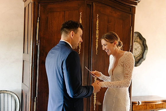 First look moment as bride reveals a photo print gift to groom in suit, wearing long sleeve lace dress in a room with vintage armoire