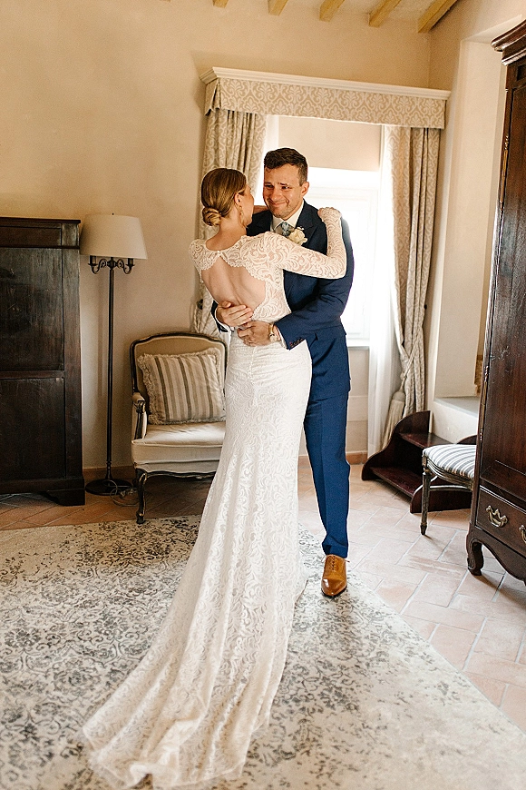 First look moment as bride hugs groom in a navy suit, her long-sleeve lace open-back gown glowing in soft window light indoors