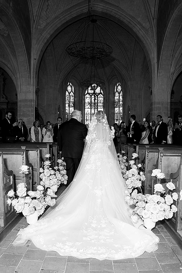 Bridal processional with bride walking down aisle in a lace dress and long veil, past white rose pots in a stained-glass stone church