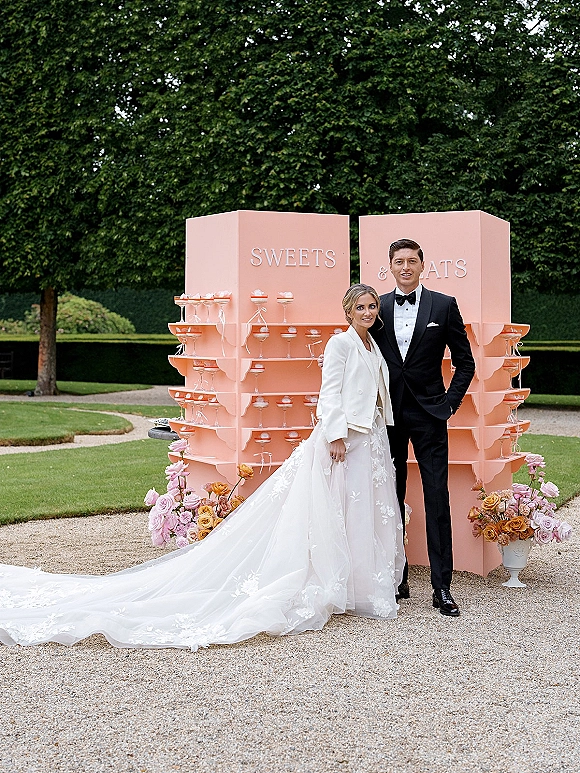 Couple portrait of bride in white coat and groom in black tuxedo beside blush dessert display with coupe glasses, roses, and hedges