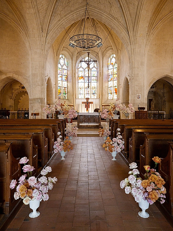 Church ceremony decor with wedding ceremony aisle flowers in pedestal urns, pink rose arrangements leading to a crucifix beneath a chandelier