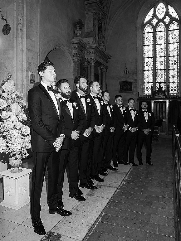 Groomsmen portrait of men lined up in black tuxedos with bow ties and boutonnieres at a church altar beneath stained glass windows