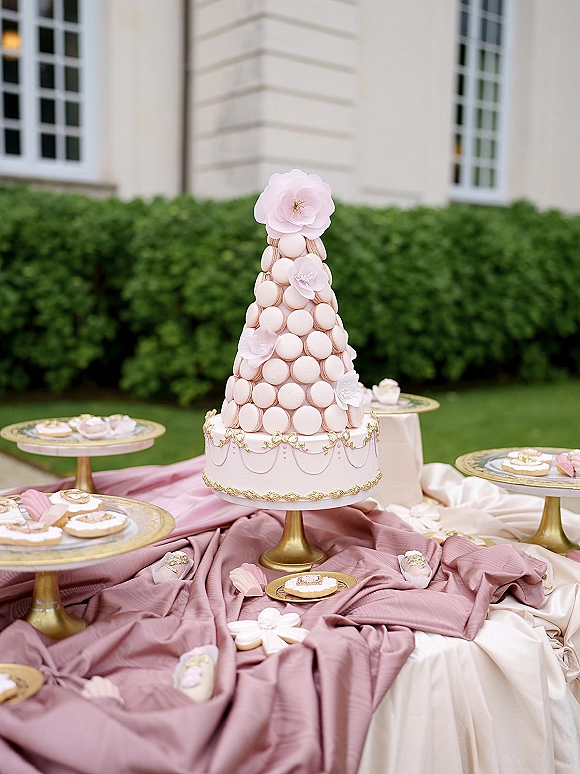 Wedding cake with macaron tower cake topper, blush frosting and sugar flowers on a gold stand, set on draped linens outdoors