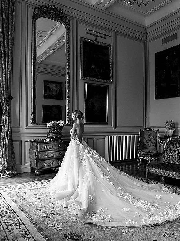 Bridal portrait of a bride in a backless gown, lace train flowing as she looks in an ornate mirror in a vintage sitting room with chandelier