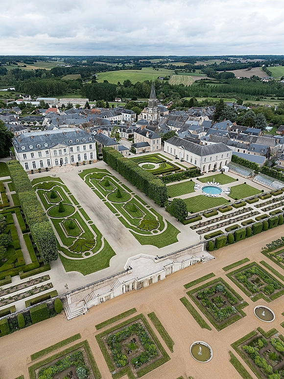 Chateau wedding venue with french chateau exterior overlooking formal parterre gardens, fountain basins, and pool on a stone terrace under clouds