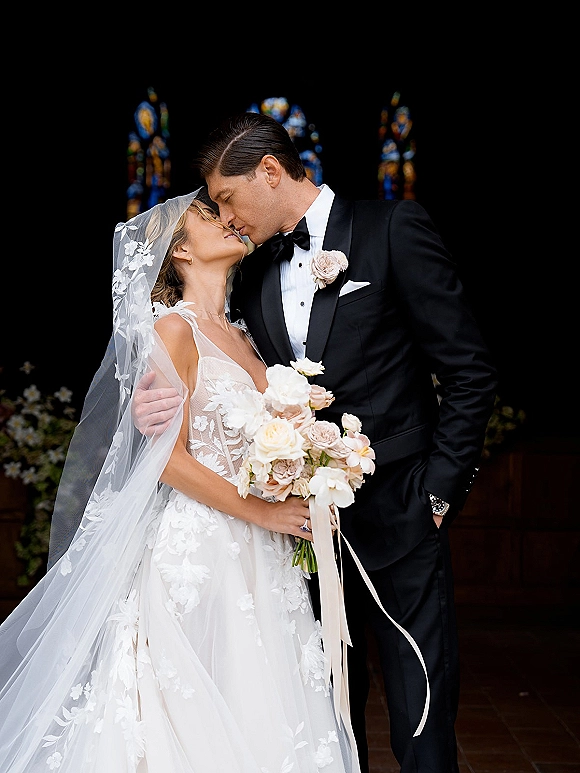 Wedding kiss portrait of bride and groom kissing in a church doorway, her cathedral veil and blush rose bouquet against stained glass windows