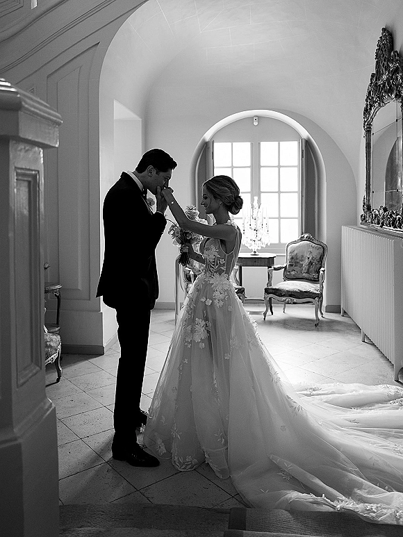 Wedding couple portrait of groom kissing the bride’s hand, her lace dress train trailing in a window-lit hallway with arched doorway and chandelier