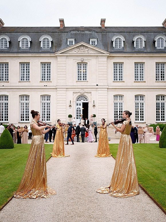 Wedding musicians in gold sequin gowns play violins as guests arrive along a chateau driveway with trimmed hedges and lawn