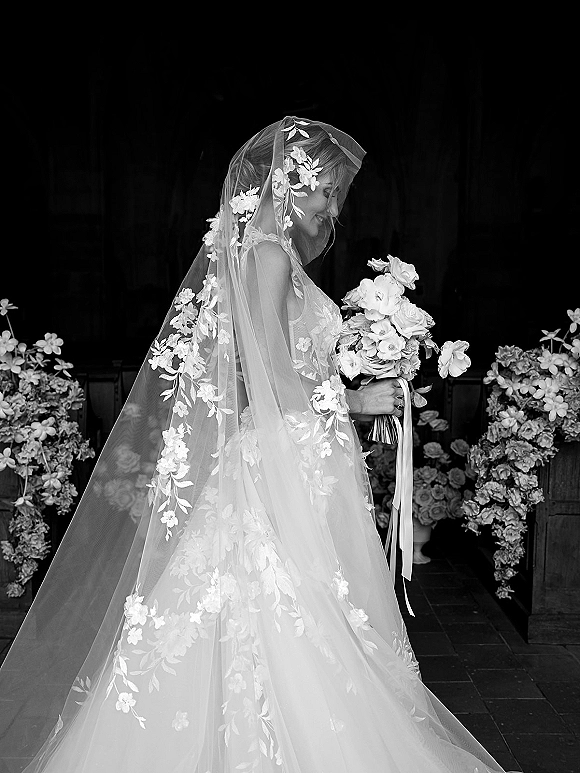 Bridal portrait of a bride holding a bouquet, smiling in side profile with a floral embroidered veil in a dark doorway backdrop