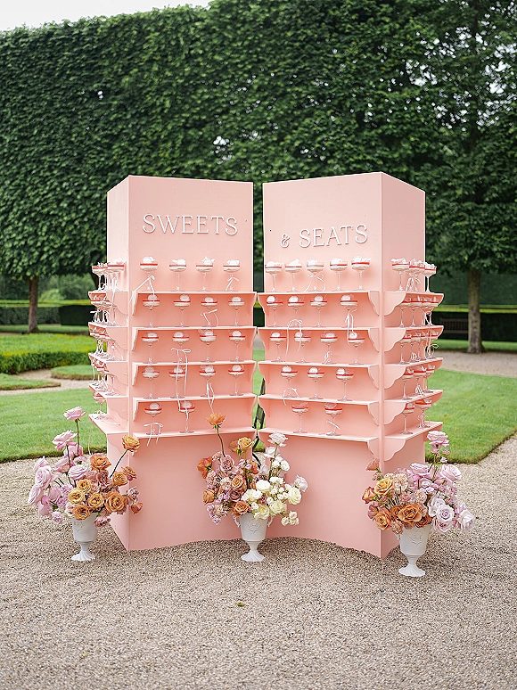 Wedding escort display with escort card display wall featuring coupe glasses, ribbons, and rose florals along hedges and gravel path outdoors