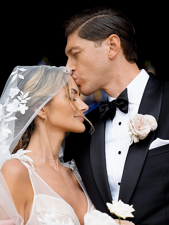 Wedding couple portrait of groom kissing the bride’s forehead, her lace veil and earrings glowing against a dark blurred background