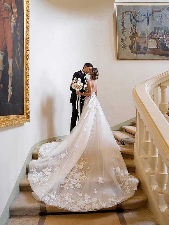 Wedding kiss portrait of bride and groom kissing on a grand staircase, bride holding a bouquet with ribbon wrap and lace train flowing