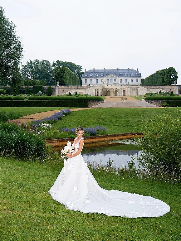 Bridal portrait of a bride holding bouquet in a long-train lace wedding dress with veil on a stone terrace by a garden pond and manor house