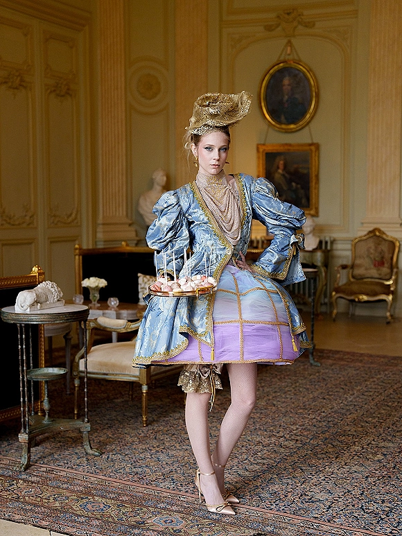 Bridal fashion portrait of a bride in a blue brocade puff-sleeve dress with gold fascinator, holding a dessert tray in an ornate room