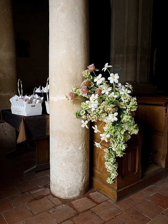 Wedding floral arrangement at ceremony entrance with white blooms, blush roses and cascading greenery on a pedestal beside petal cones near a doorway