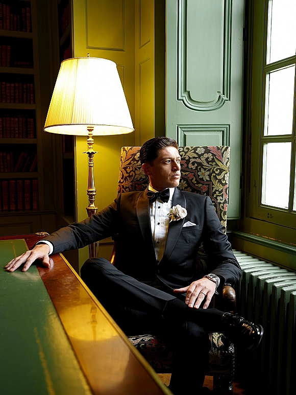 Groom portrait in a black tie tuxedo with boutonniere, bow tie, and wristwatch, seated by window light in a paneled study room