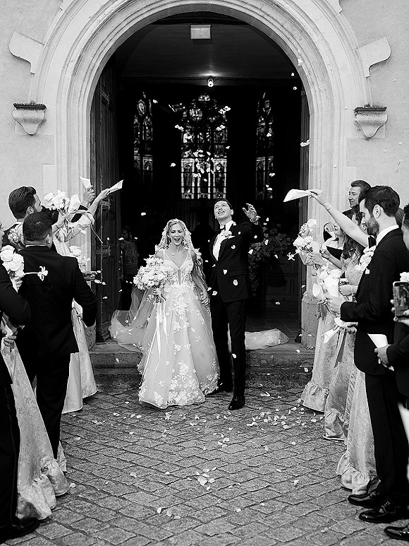 Wedding recessional as bride and groom walk out of a church wedding exit, holding hands under rose petals and confetti at stone arch doorway