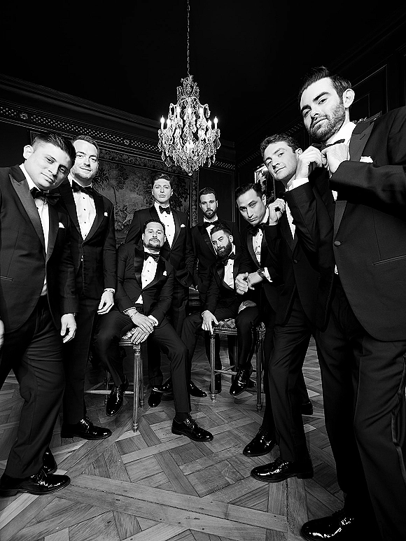 Groomsmen portrait of men in black tuxedos with bow ties and boutonnieres posing in an ornate room with dark paneled walls and chandelier