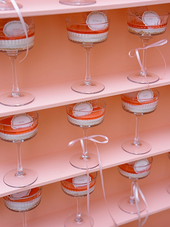 Wedding escort display of cocktail glass escort cards with layered gelatin desserts in coupe glasses, satin ribbon bows, on tiered shelves against a peach backdrop