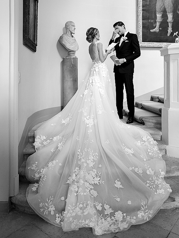 Wedding vows as bride in a long-train, floral applique gown and groom in tux read from booklets on an indoor staircase with paintings