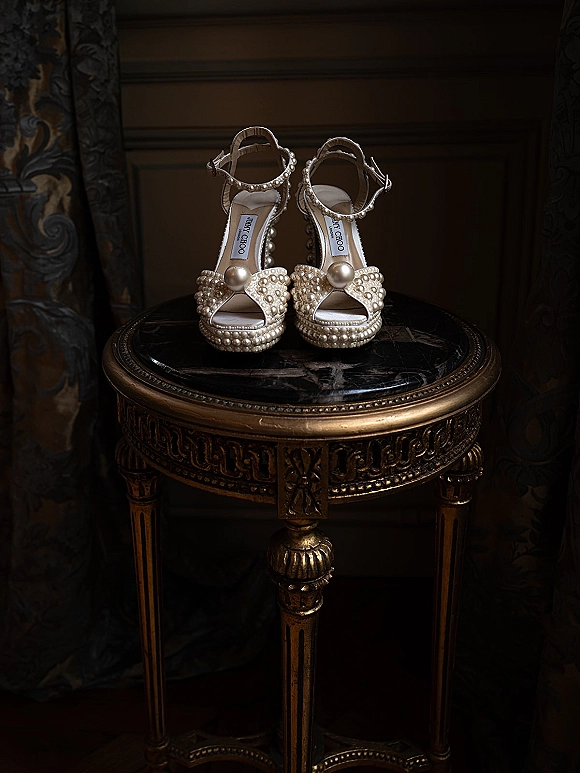 Bridal shoes with pearl wedding shoes details, ankle straps and bow on a marble tabletop beside a gilded table in a moody room