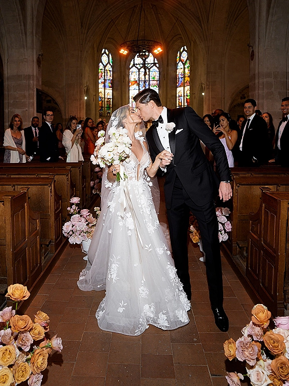 Wedding kiss as bride and groom walk down the aisle, her cathedral veil and bouquet flowing past pews and stained glass windows
