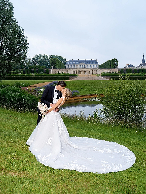 Wedding kiss portrait of bride and groom kissing in a dip, bride holding bouquet with lace train, groom in tuxedo on estate garden lawn by pond