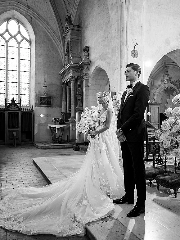 Ceremony moment at a church wedding ceremony as bride in lace veil and long train stands with groom in tuxedo at the altar