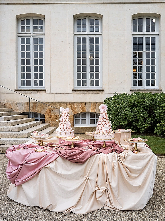Wedding dessert table with macaron tower wedding display, blush and ivory draping, gold cake stands, set in a courtyard by tall windows