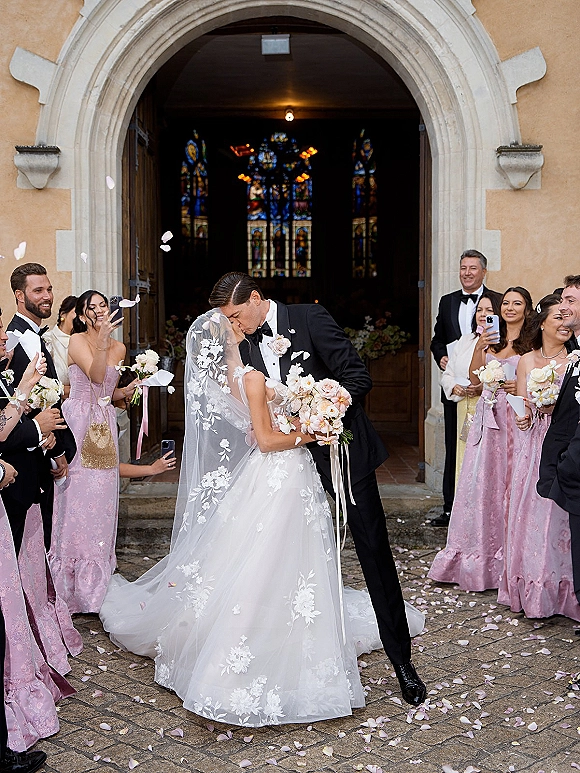 Wedding kiss portrait as bride dips in lace dress and veil, groom in tuxedo, while petals fly and guests film at church doorway stone arch