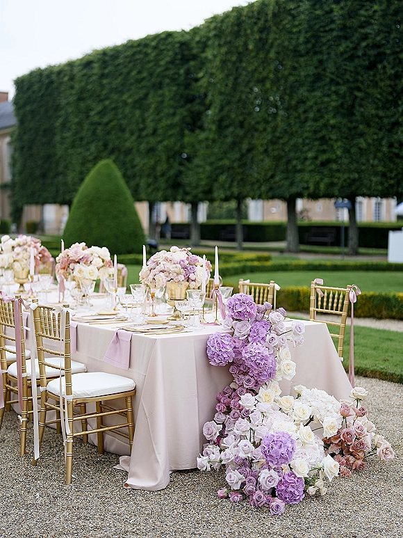 Reception tablescape with an outdoor reception table of garden roses and hydrangeas, gold compote vases, taper candles, and lavender napkins on a gravel patio