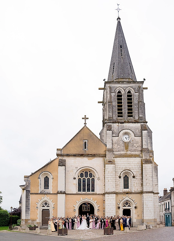 Wedding group photo of bride and groom with guests outside a stone church, bridesmaids in pink and navy-suited groomsmen with bouquets under overcast sky