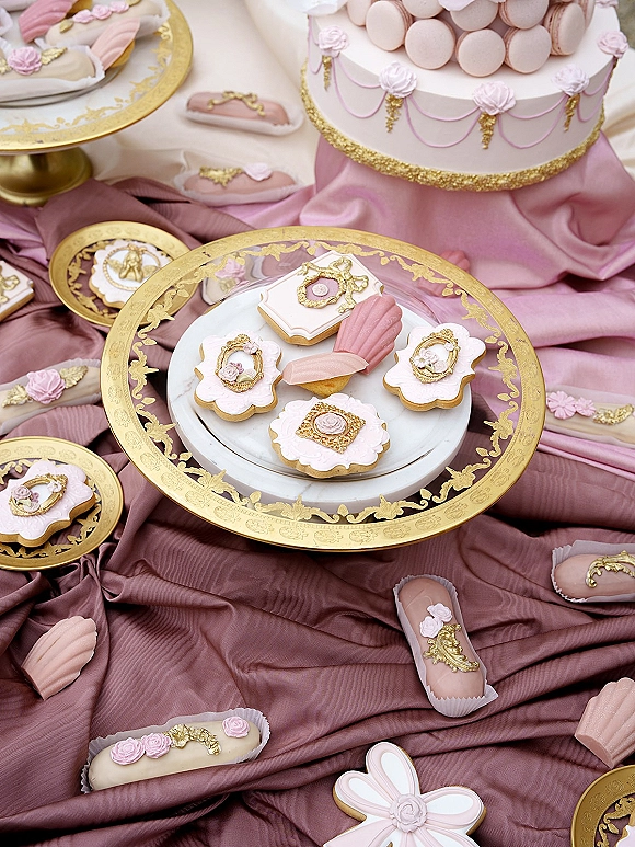 Wedding dessert table featuring wedding sugar cookies, macarons, and a pink fondant cake with gold trim on patterned plates before draped fabric backdrop