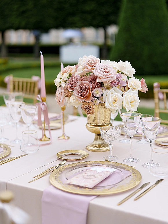 Reception tablescape with an outdoor reception table set in pink and gold, featuring a rose centerpiece in a gold compote on a garden lawn