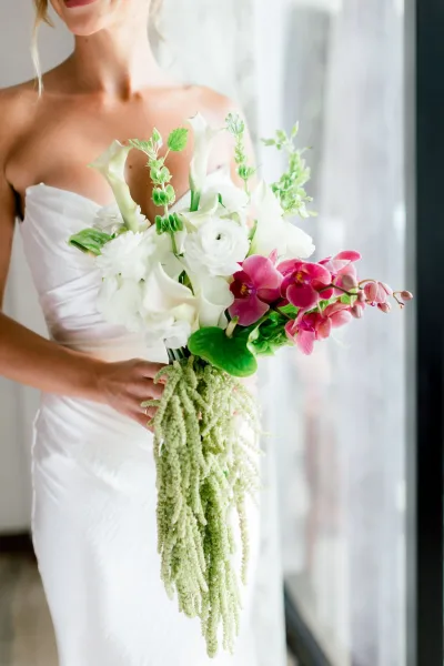 Bridal bouquet of white calla lilies and pink orchids with trailing amaranthus held against a strapless dress by window light indoors