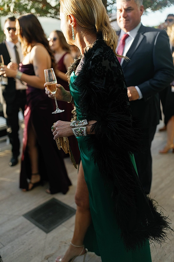 Wedding guest style in an emerald green satin dress with black feather shawl, holding champagne on a sunlit patio among mingling guests