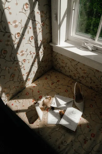 Wedding invitation suite flat lay with envelopes, RSVP card, rings, and bridal shoes tied with ribbon and wax seal in window sunlight