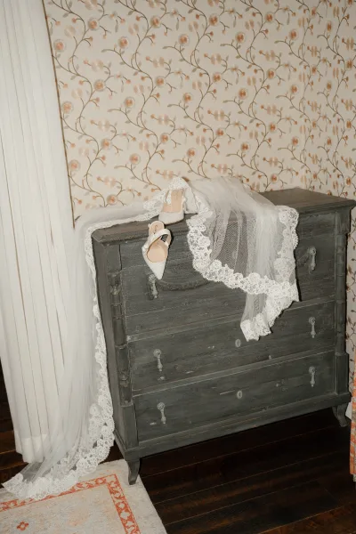 Bridal accessories with lace wedding veil draped beside white heels on a vintage dresser, set against floral wallpaper and hardwood floor