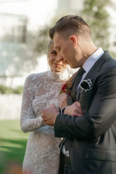 Couple portrait of bride looking at groom in a black tuxedo with red boutonniere, her long-sleeve lace high-neck dress in sunlit greenery