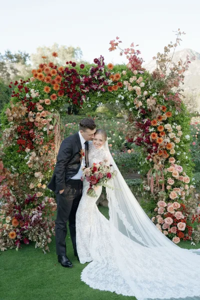 Couple portrait of bride and groom embrace as he kisses her forehead, lace gown and long veil under a floral arch with mountains behind