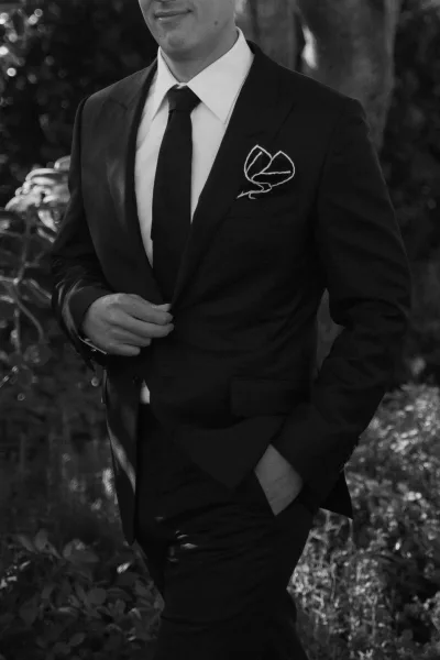 Groom portrait in a black tie suit with white dress shirt and pocket square, posed with hands in pockets amid garden foliage and trees