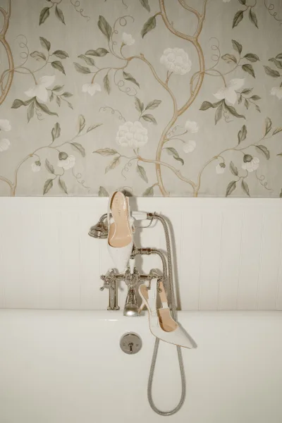 Bridal shoes with rhinestone sparkle and ankle straps perched on a bathtub faucet, framed by white wainscoting and floral vine wallpaper