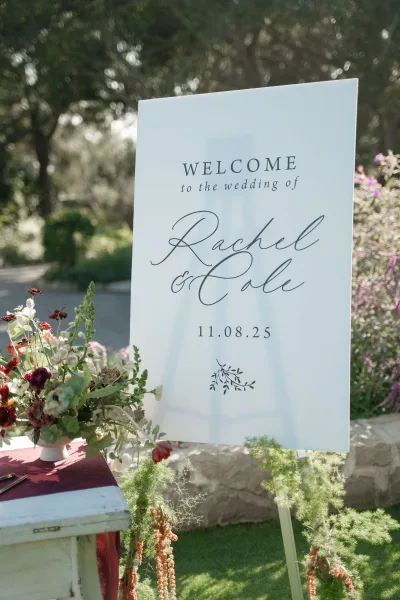 Wedding welcome sign with calligraphy on a white acrylic panel displayed on an easel, styled with greenery and florals in a garden path setting