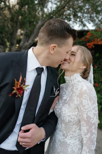 Wedding kiss portrait of bride and groom kiss, her long-sleeve high-neck lace dress and veil against lush garden greenery and trees