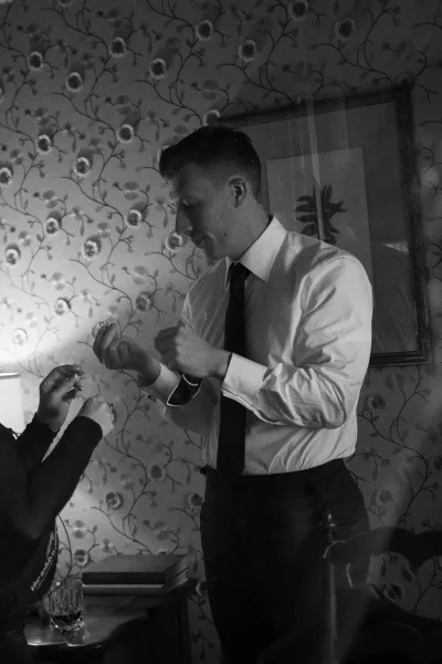 Groom getting ready, putting on cufflinks with a wristwatch and ring beside him in a room with floral wallpaper and framed artwork