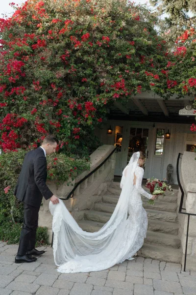 Couple portrait of bride and groom walking up stone steps, groom lifting her lace dress train beneath red bougainvillea vines