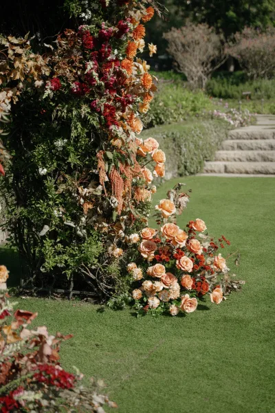 Floral ceremony arch with roses, dahlias, and hanging amaranthus in rust and peach tones on a garden lawn near stone steps