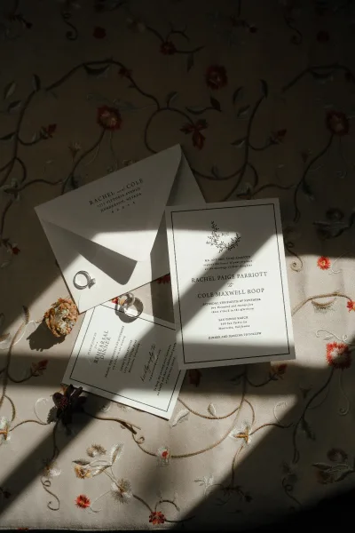 Invitation suite wedding invitation flat lay with envelope, RSVP card, rings, wax seal, and dried flowers on embroidered floral fabric in sunlight shadows